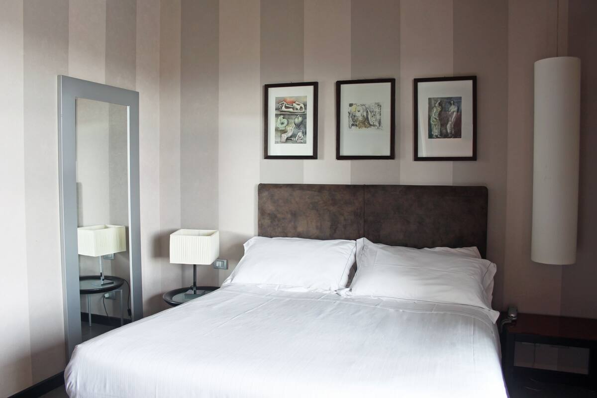 A hotel room with a large bed, a bedside table with a lamp, a mirror, and three framed artworks on the wall.