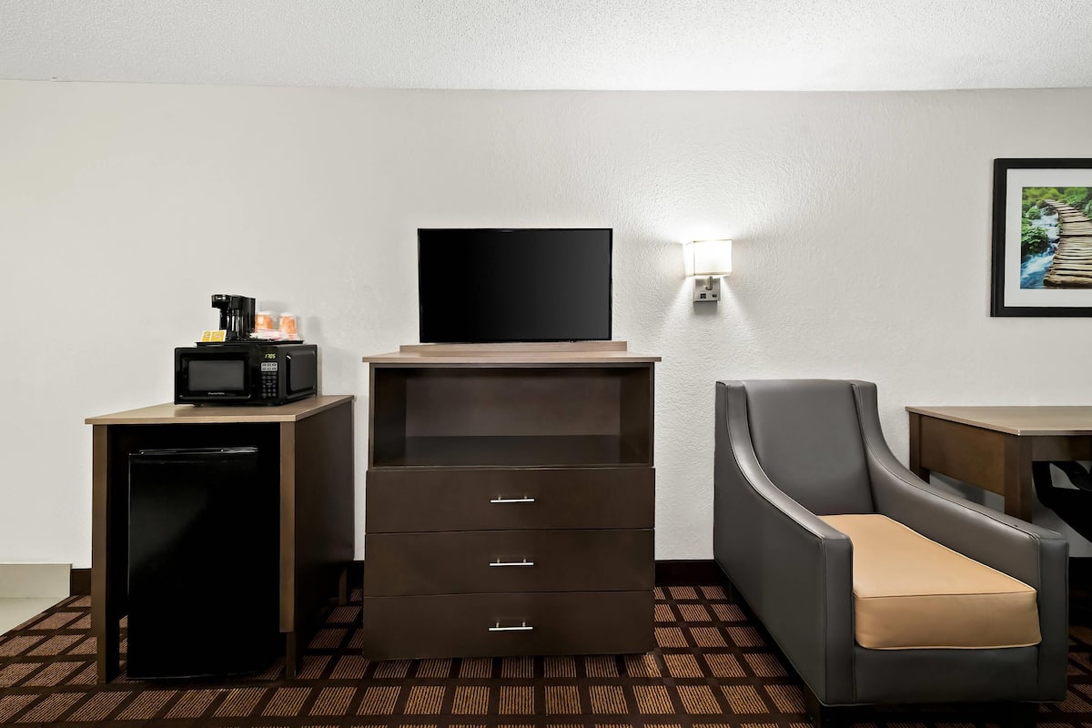 Ein Hotelzimmer mit Flachbildfernseher auf einem Holzständer, einem Schreibtisch mit Mikrowelle, einem Kühlschrank, einem Sessel und einer Wandlampe.