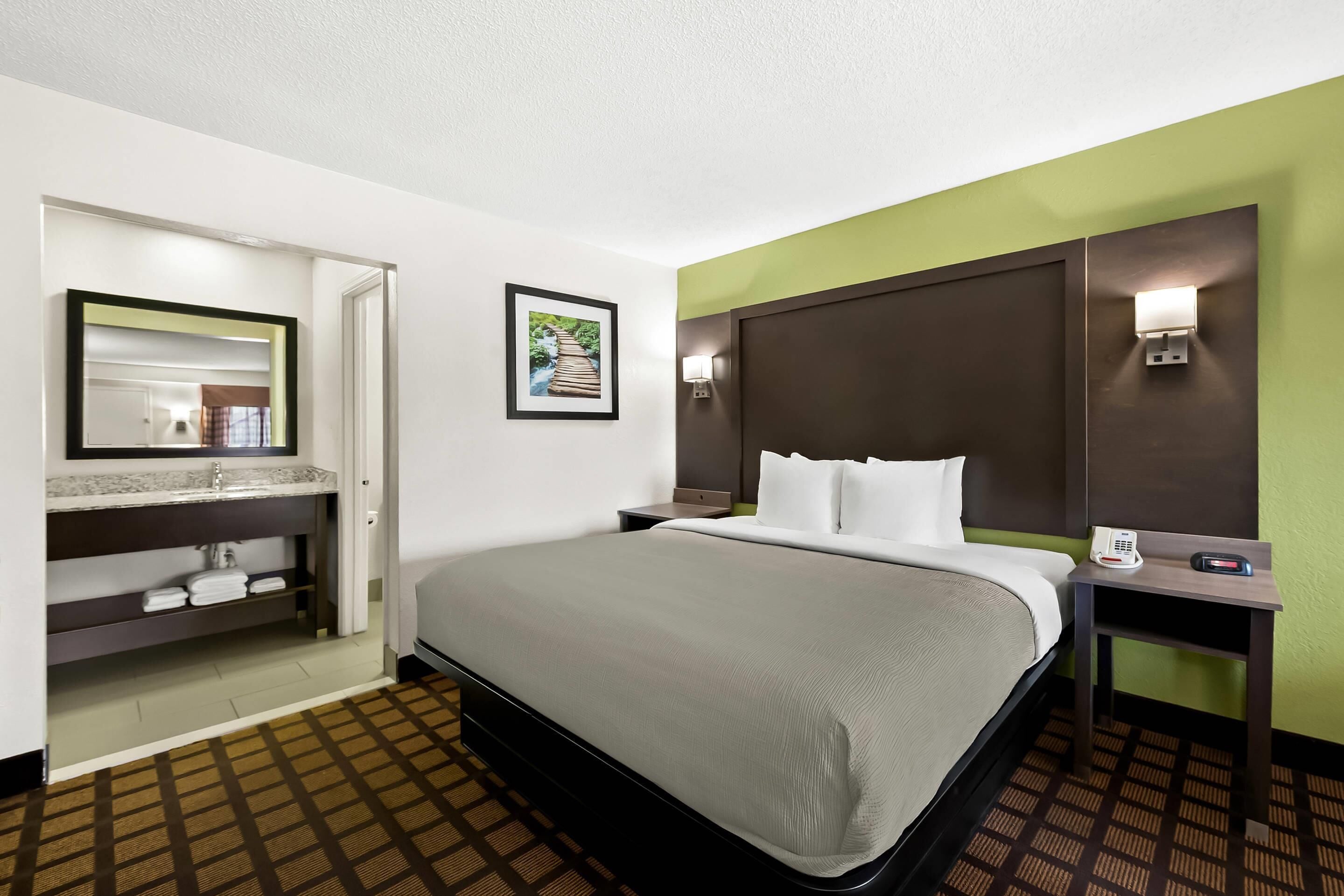A hotel room with a bed, a bathroom with a mirror, a picture on the wall, and a nightstand with a phone.