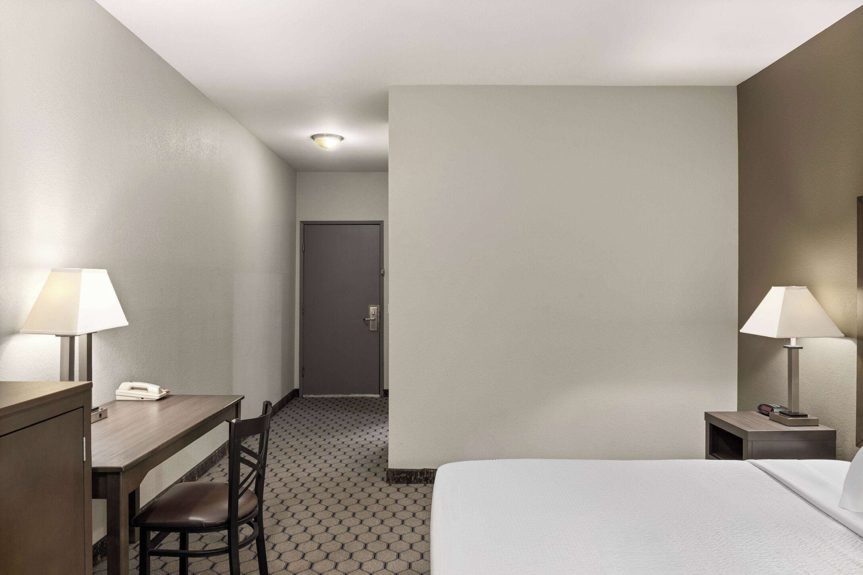 A hotel room with a bed, a desk with a telephone, a chair, two bedside lamps, and a door leading to another room.