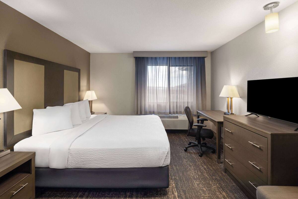 A hotel room with a large bed, two bedside tables with lamps, a desk with a chair, a television, and a large window with curtains.