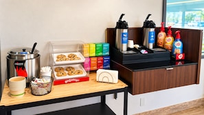 Una estación de desayuno autoservicio con una máquina de café, dispensadores de té y café, una bandeja de galletas y una variedad de opciones para el desayuno.