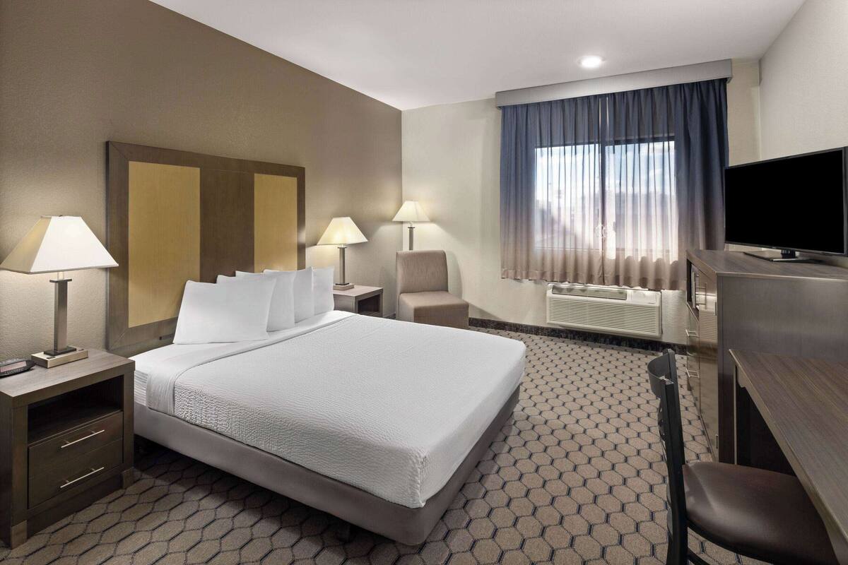 A hotel room with a large bed, two bedside tables with lamps, a TV, a desk with a chair, and a window with curtains.