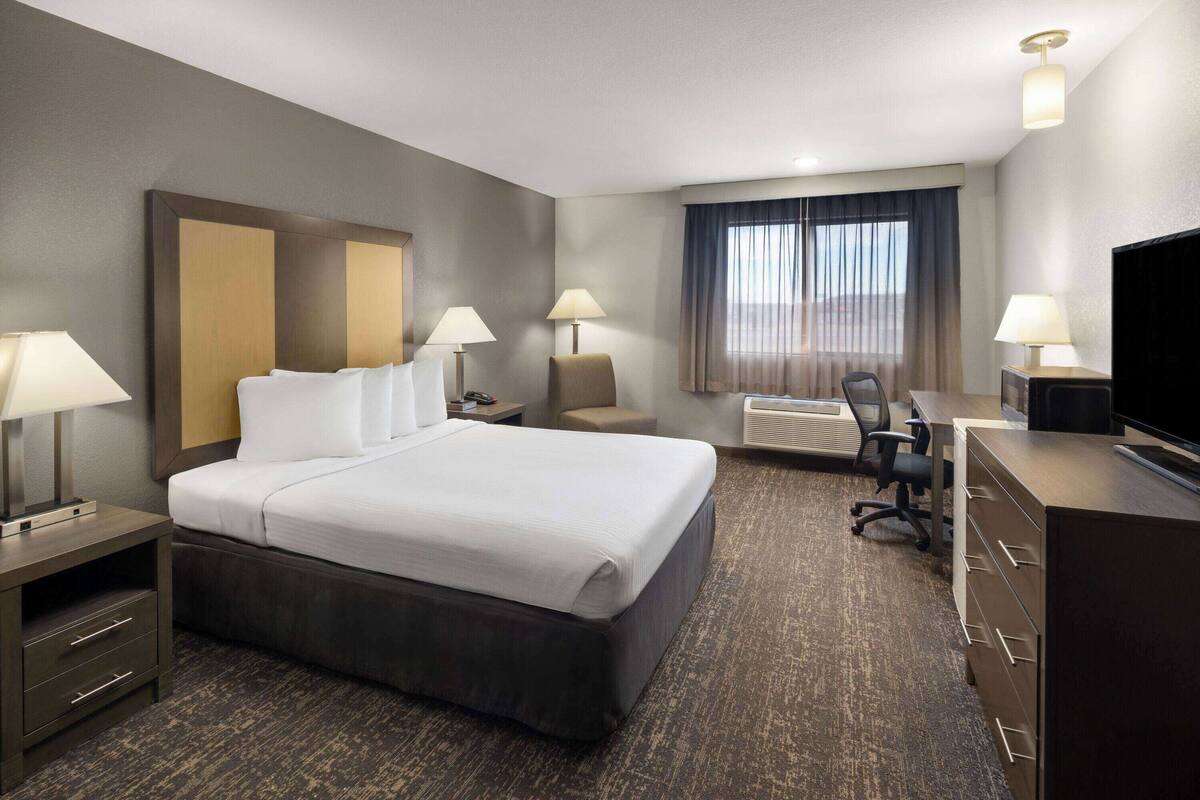 A hotel room with a large bed, two bedside tables with lamps, a desk with a chair, a television, and a window with curtains.