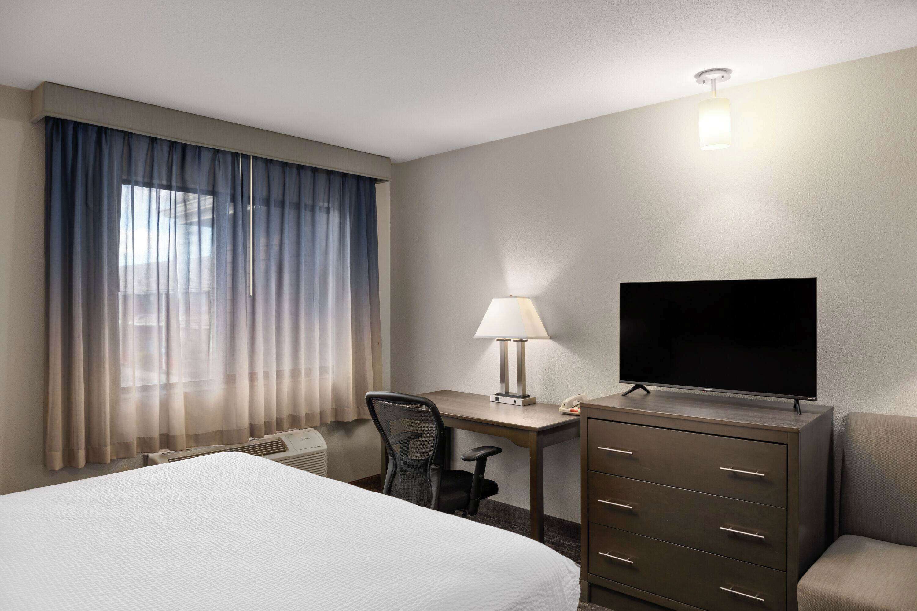 A hotel room with a bed, a desk with a lamp, a television, a chair, and a dresser.