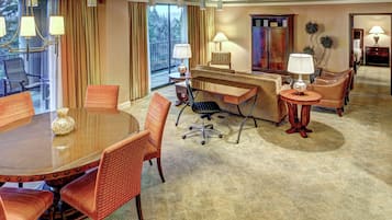 A hotel room with a large round table, several chairs, a desk with a chair, lamps, a cabinet, and a view of the outdoors.