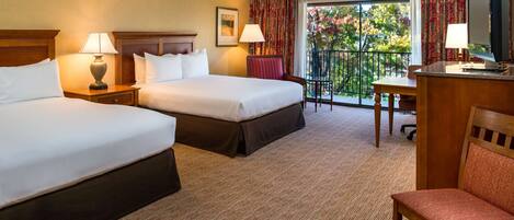 A hotel room with two beds, a desk, a chair, a TV, and a balcony with a view of trees.
