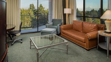 Habitación de hotel con balcón, sofá de cuero, mesa de centro de vidrio, escritorio con silla y lámpara.