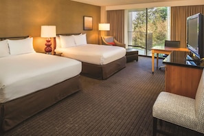 A hotel room with two beds, a desk with a television, and a balcony with a view of trees.