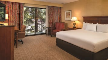 A hotel room with a large bed, a desk, a chair, a small table, a lamp, and a view of the outdoors through a sliding door.