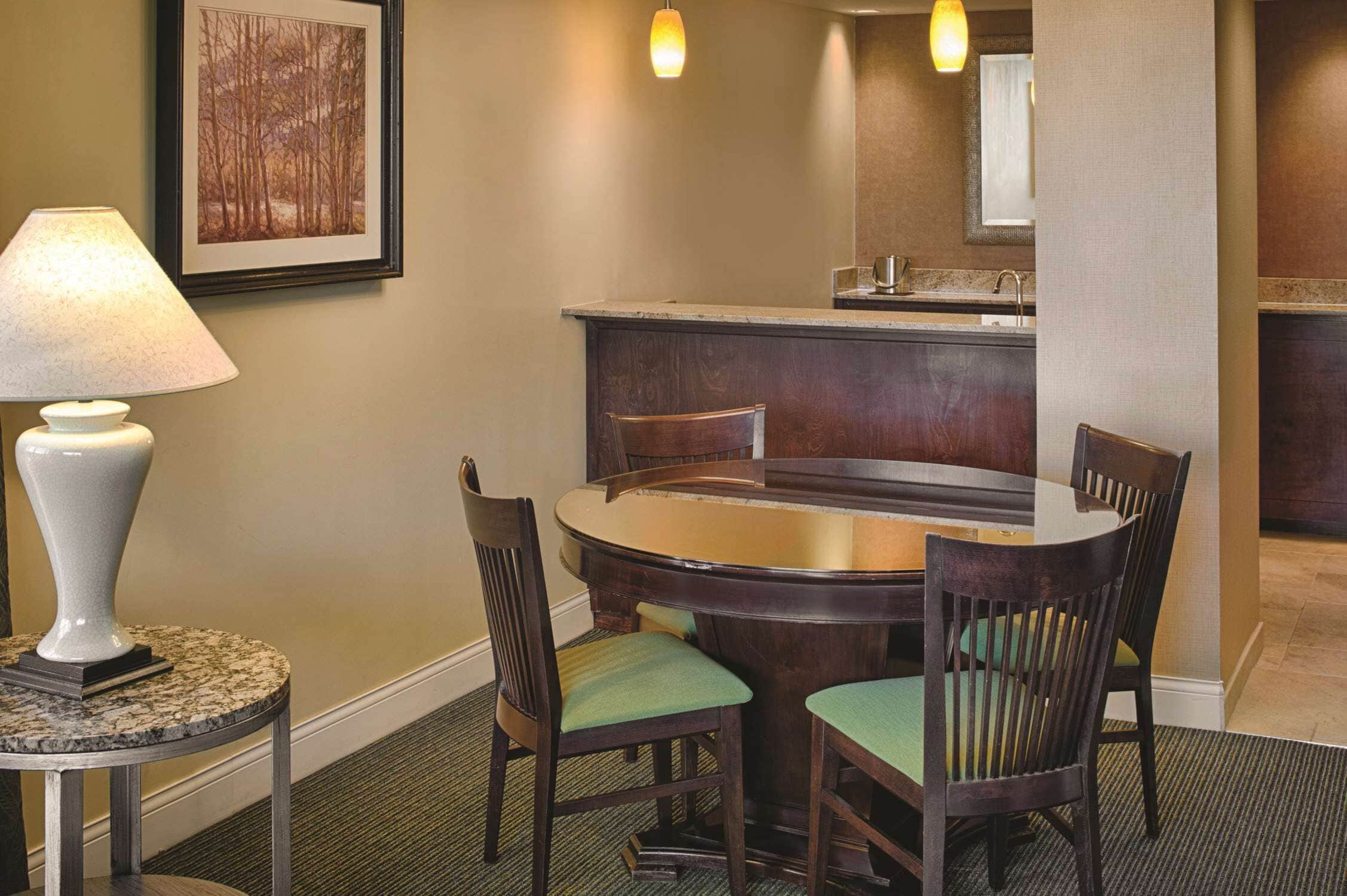 A dining area with a round table, four chairs, a lamp, and a framed picture on the wall.