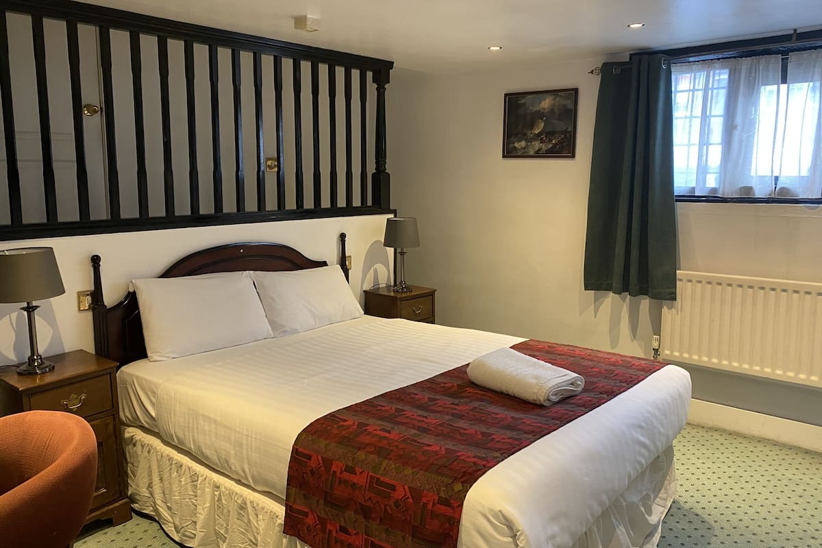 A neatly made bed with a red and white patterned blanket, a wooden headboard, a nightstand with a lamp, and a window with curtains.