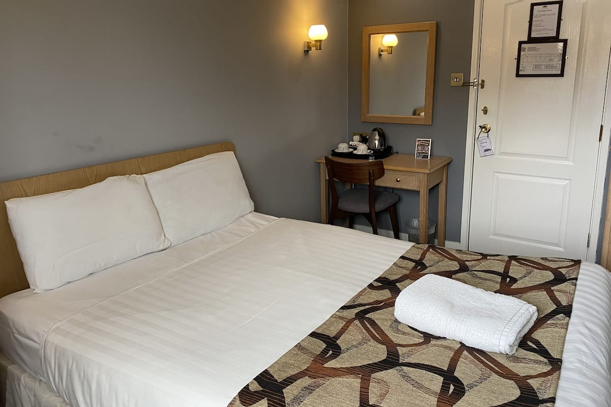 A hotel room with a bed, a desk, a chair, a mirror, and a door with a sign.