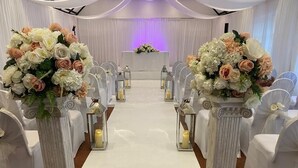 An aisle with white chairs and a white carpet, flanked by white columns with floral arrangements. - The George Hotel (Reading)