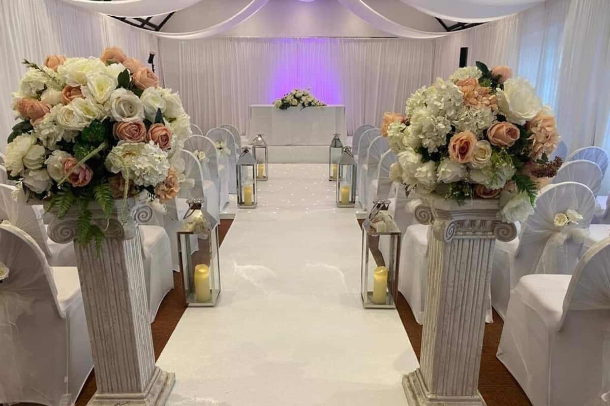 An aisle with white chairs and a white carpet, flanked by white columns with floral arrangements.