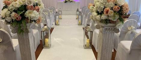 Une allée bordée de colonnes blanches ornées de compositions florales, avec des chaises blanches et un tapis blanc de chaque côté.