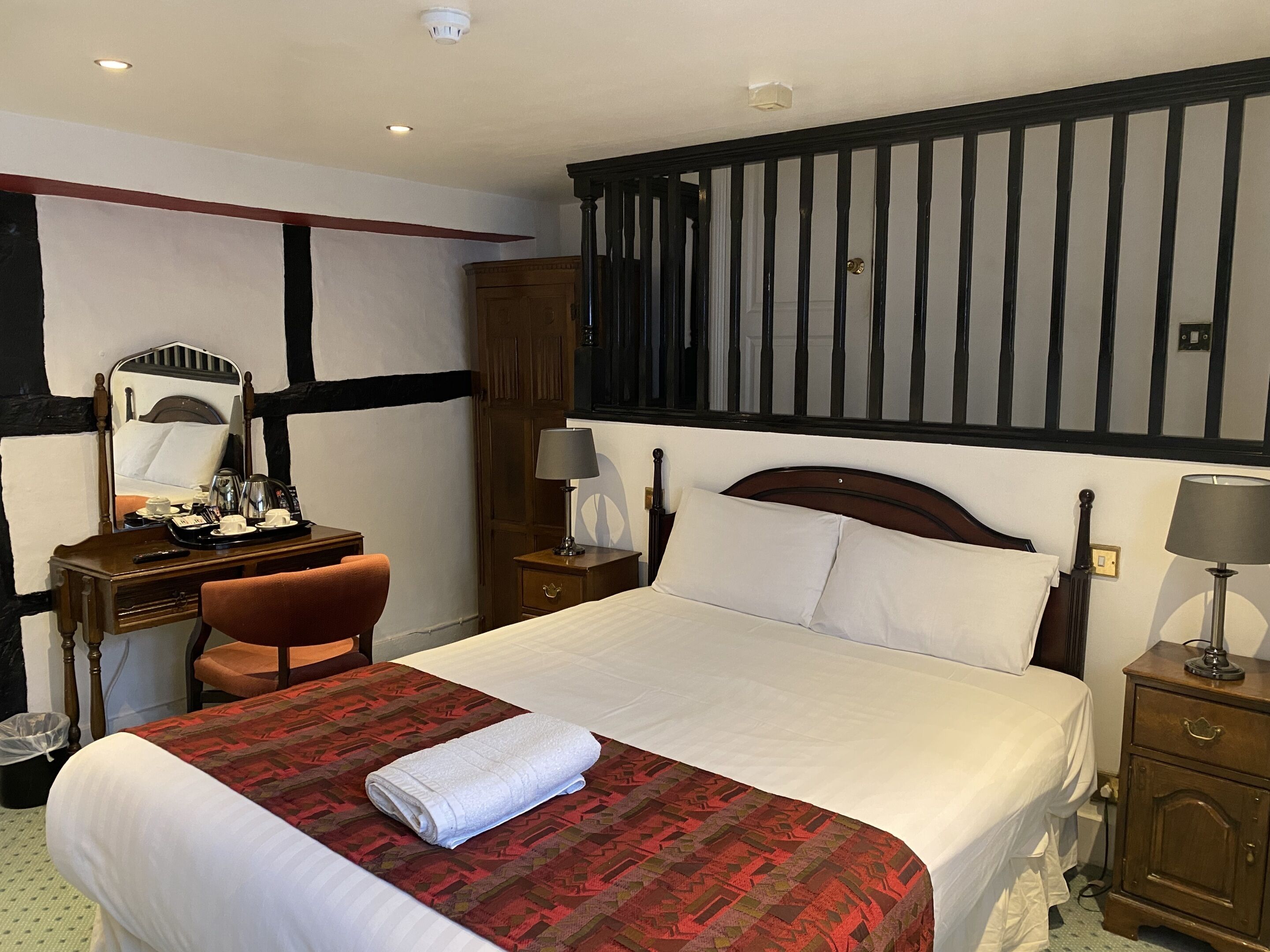 A hotel room with a large bed, a desk with a chair, a dresser, a mirror, and a nightstand with a lamp.