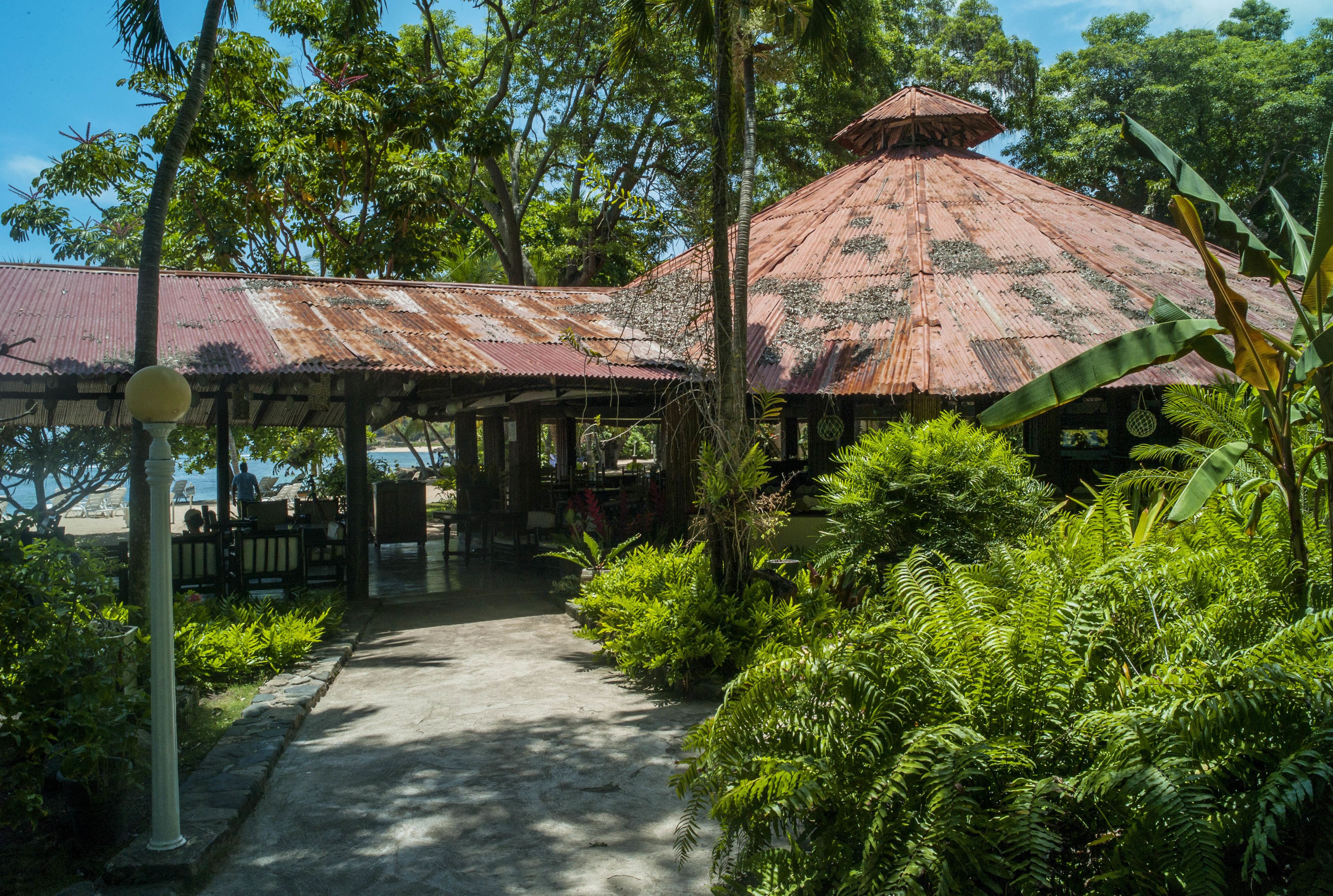 A rustic wooden structure with a thatched roof, surrounded by lush greenery and a clear view of the sea.