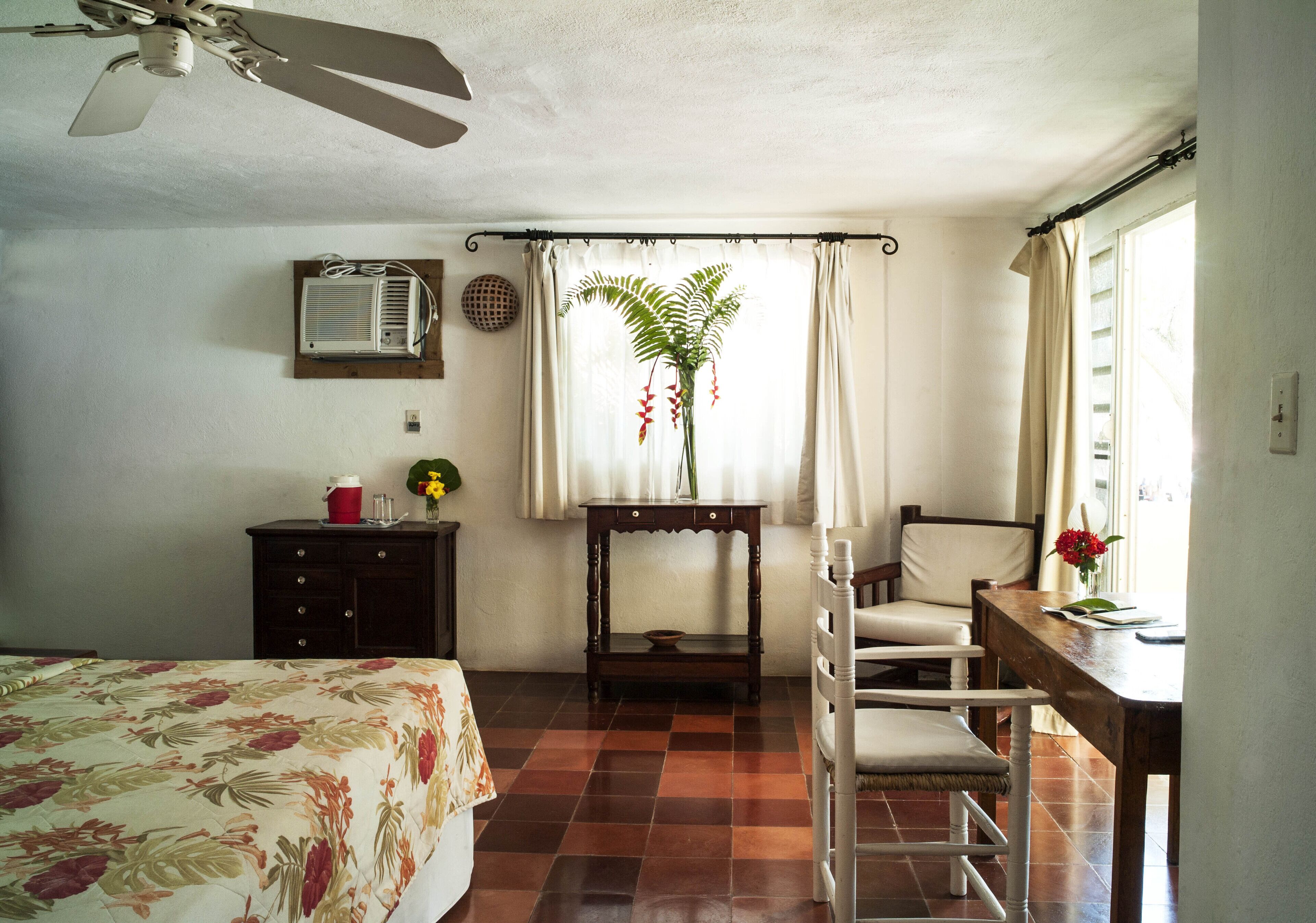 Un dormitorio con cama, escritorio, silla, ventilador de techo, aire acondicionado y ventana con cortinas.