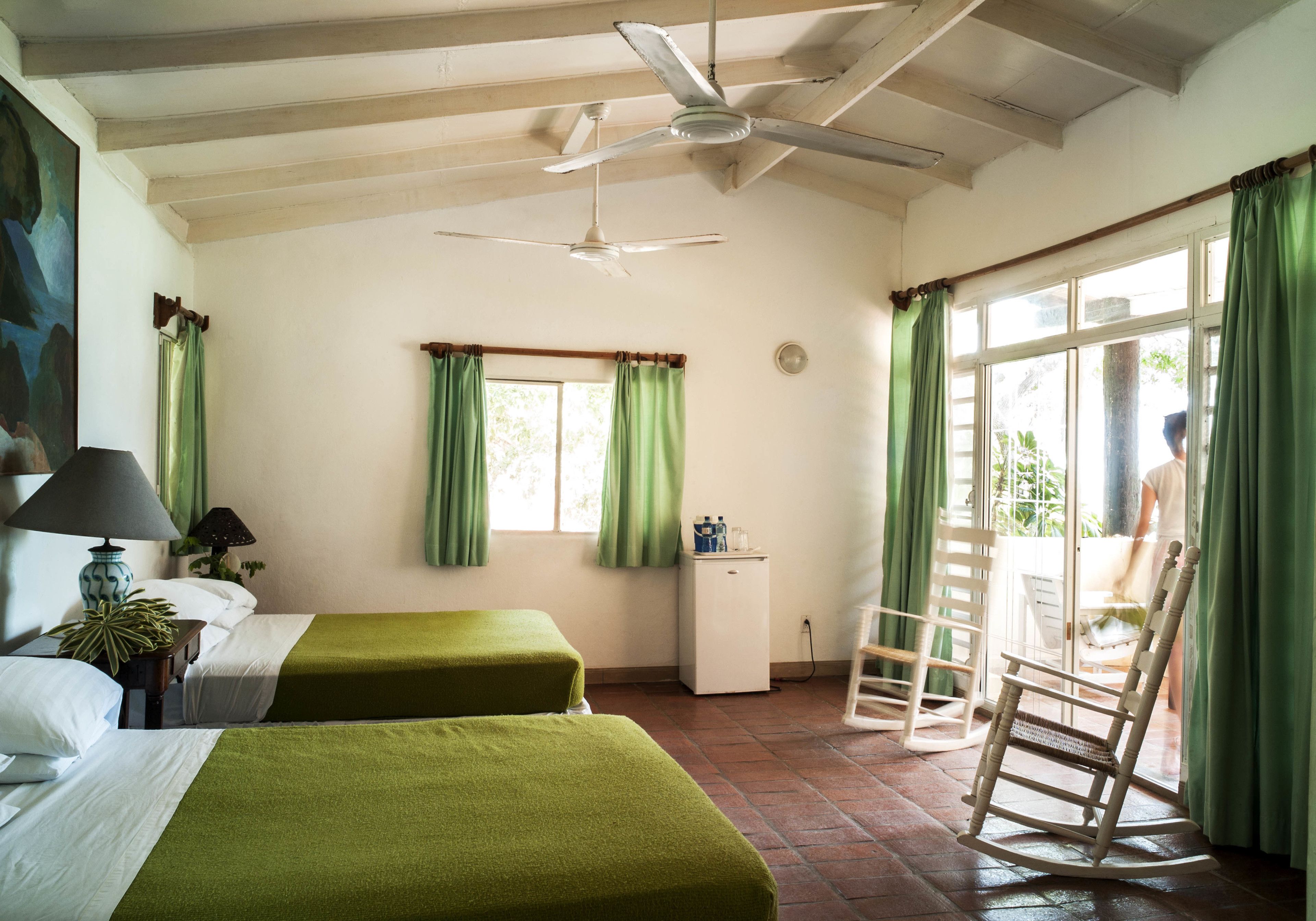 Une chambre avec deux lits, un ventilateur de plafond, une petite armoire blanche et un fauteuil à bascule.