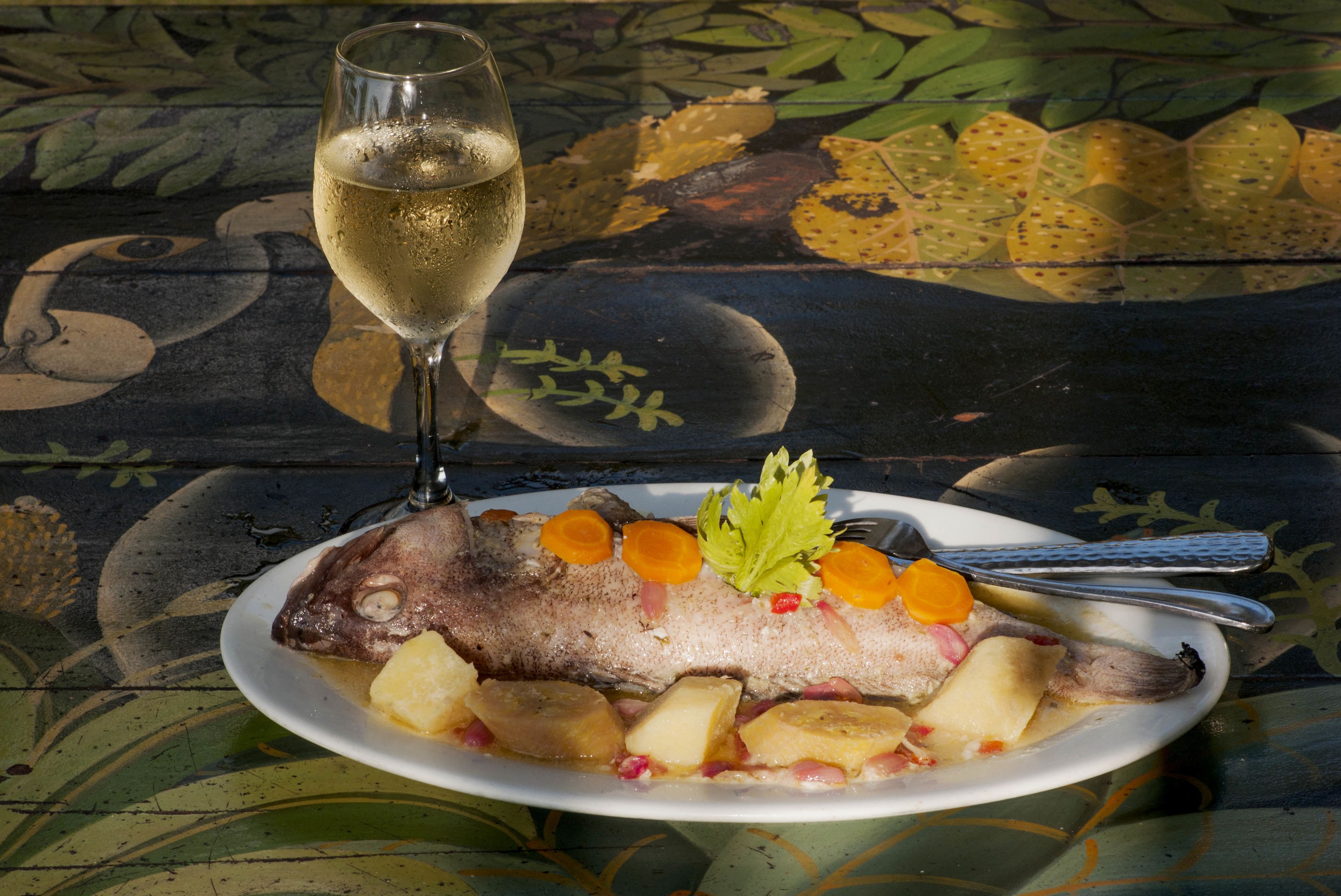 Un plato de pescado con papas y una copa de vino blanco sobre una mesa con mantel estampado con hojas.