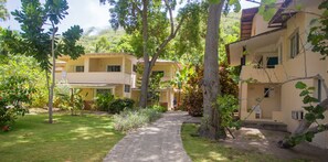 A pathway leading to a building with multiple balconies and a garden area with trees and shrubs.