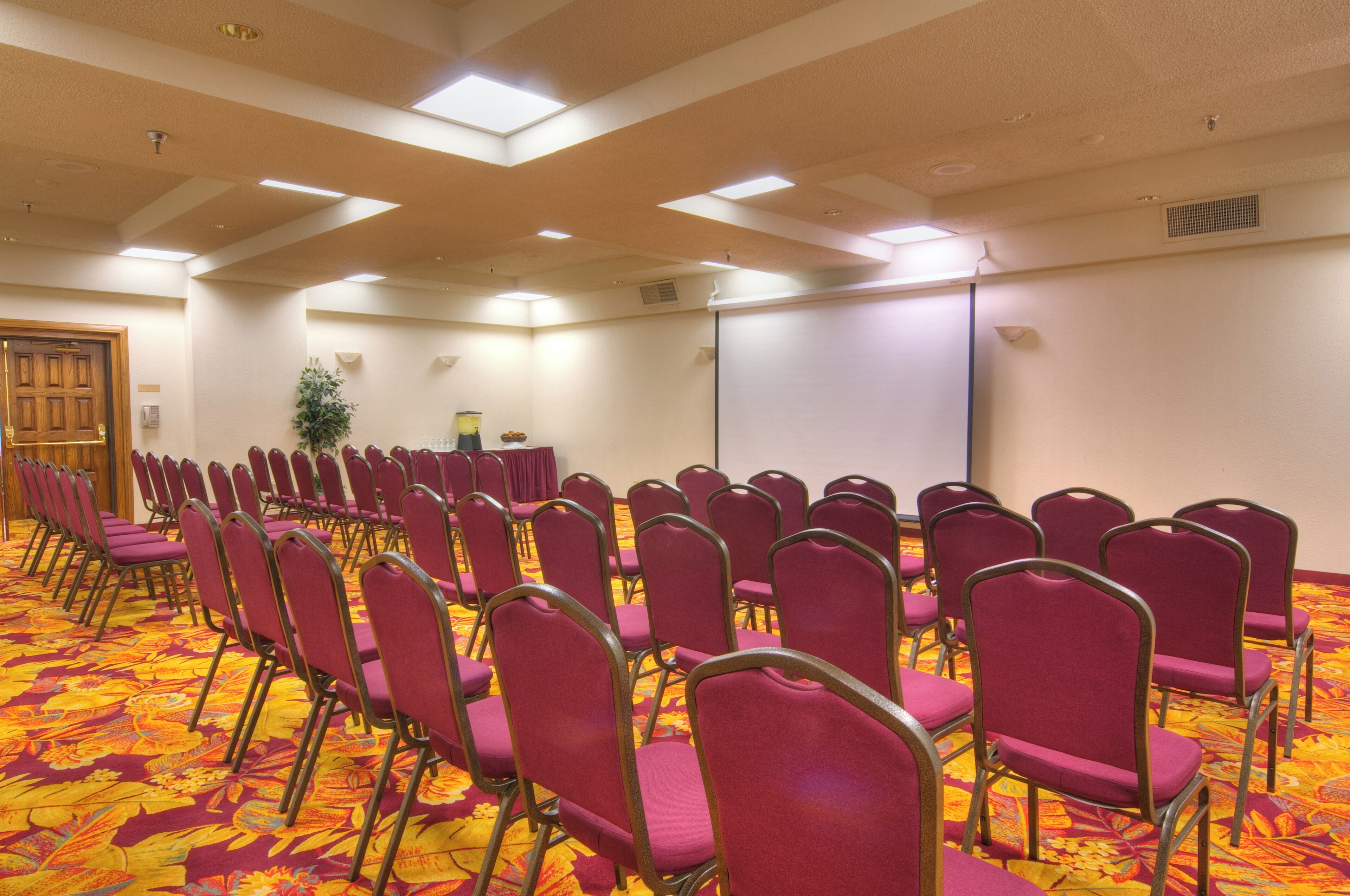 Un salón de conferencias con filas de sillas moradas, una pantalla de proyector y suelo alfombrado.