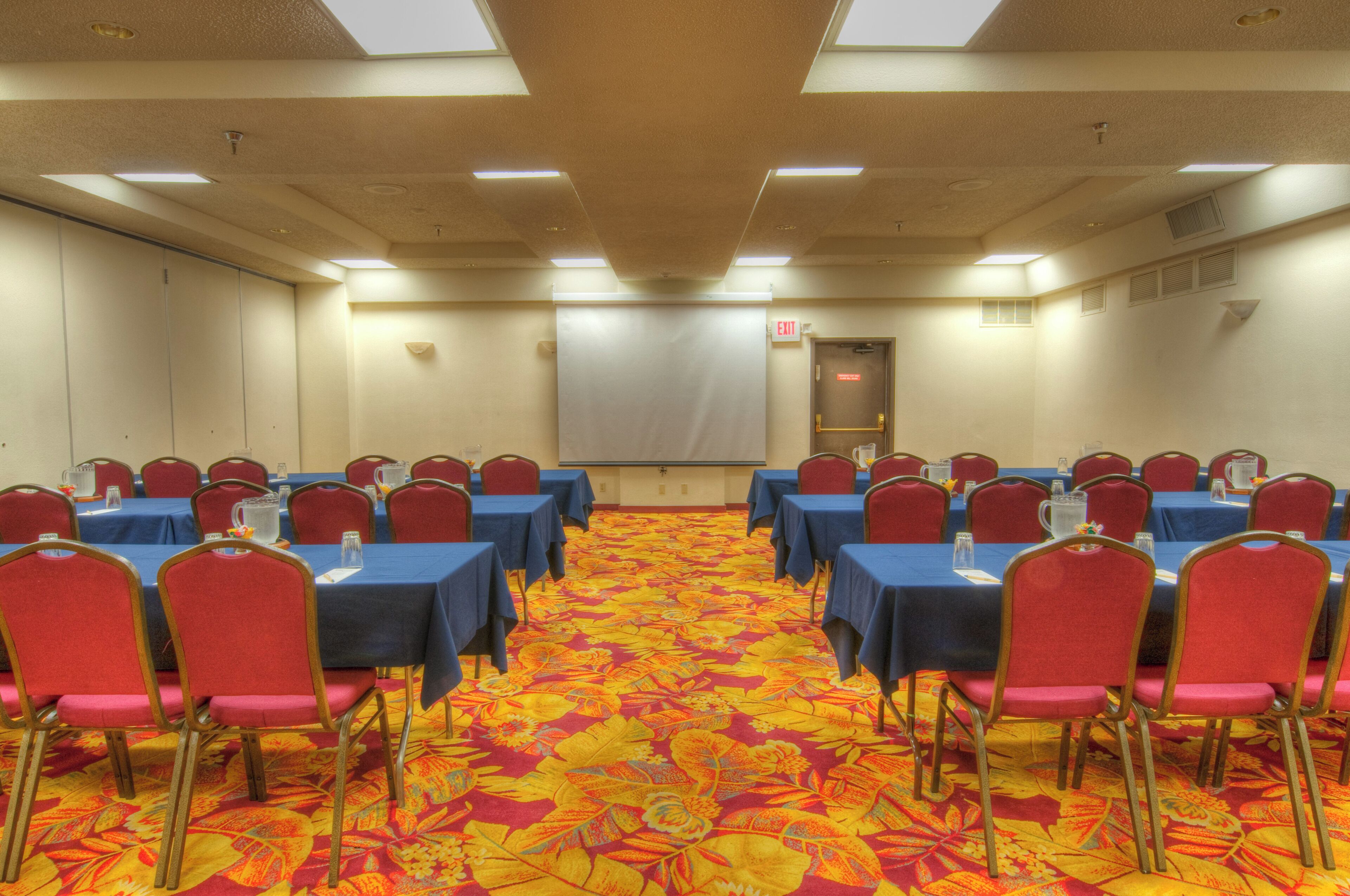 Une salle de conférence avec des rangées de tables et de chaises, un sol recouvert de moquette et un écran de projection.