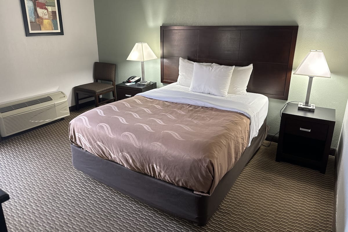 A hotel room with a large bed, two bedside tables, a nightstand, a wall-mounted air conditioner, and a painting on the wall.