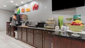 Zona de desayuno de hotel con una máquina de café, exhibición de frutas y un televisor montado en la pared.