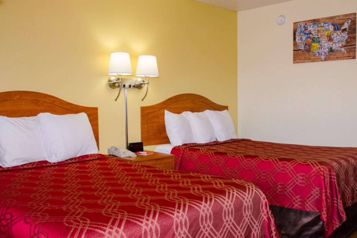 A hotel room with two beds, each with a red patterned bedspread and white pillows. There is a nightstand with a phone and a lamp between the beds.
