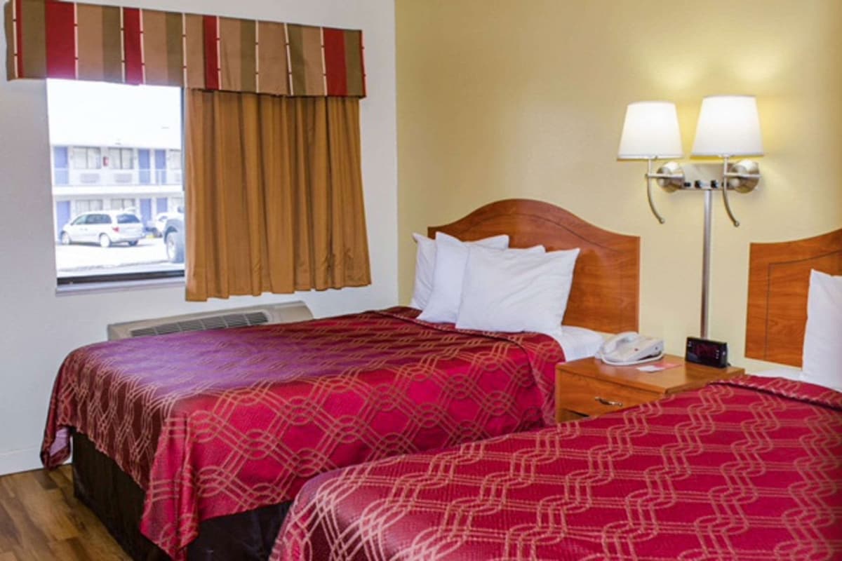 A hotel room with two beds, a window with curtains, a wall-mounted lamp, and a view of a building outside.