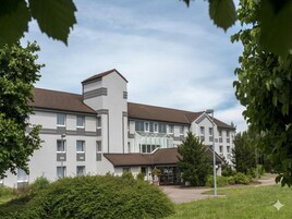 A multi-story hotel with a modern design, featuring a central entrance with glass doors, surrounded by greenery and a paved driveway.