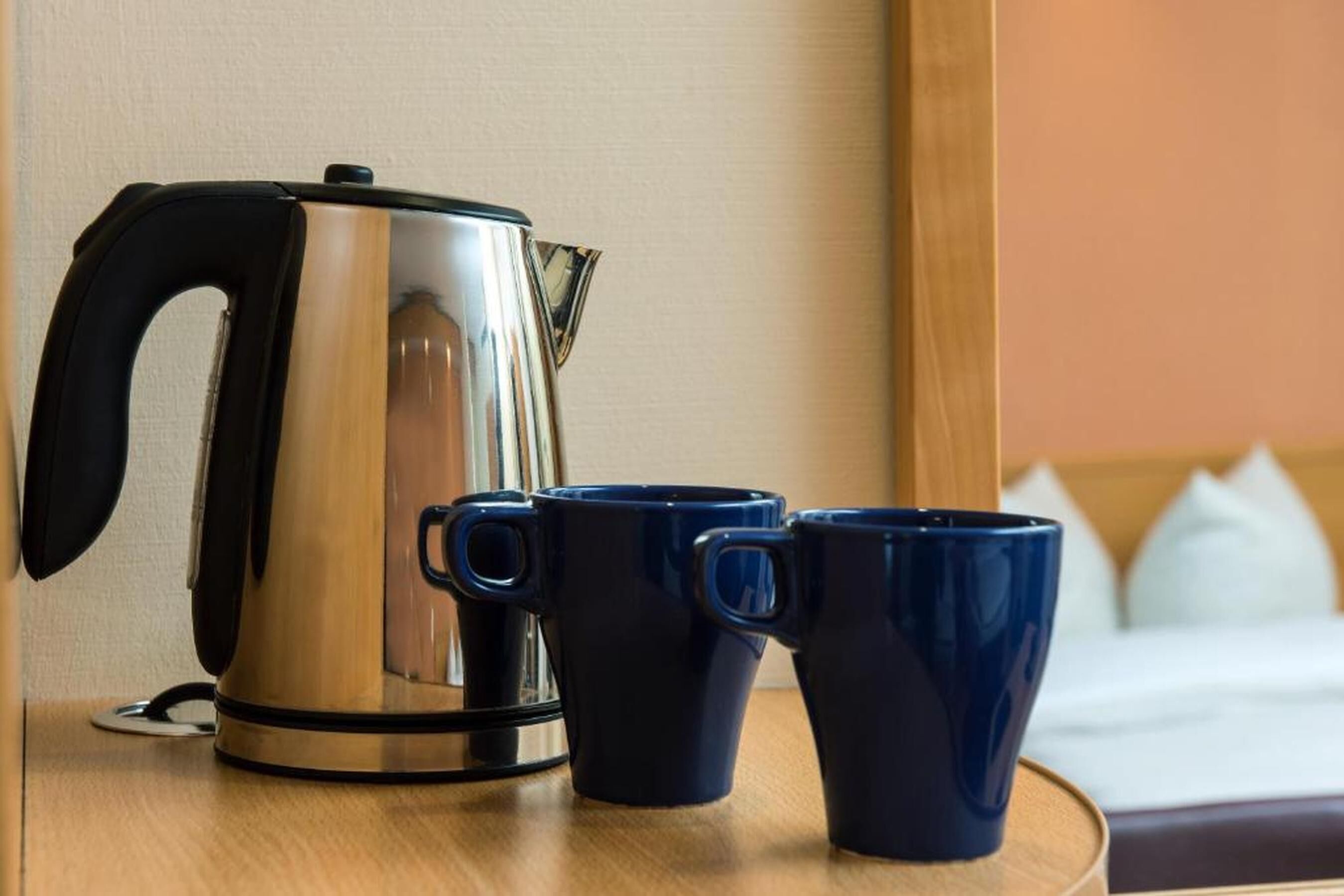 Une bouilloire en acier inoxydable et deux tasses bleu foncé sur une table en bois, dans une chambre d’hôtel.