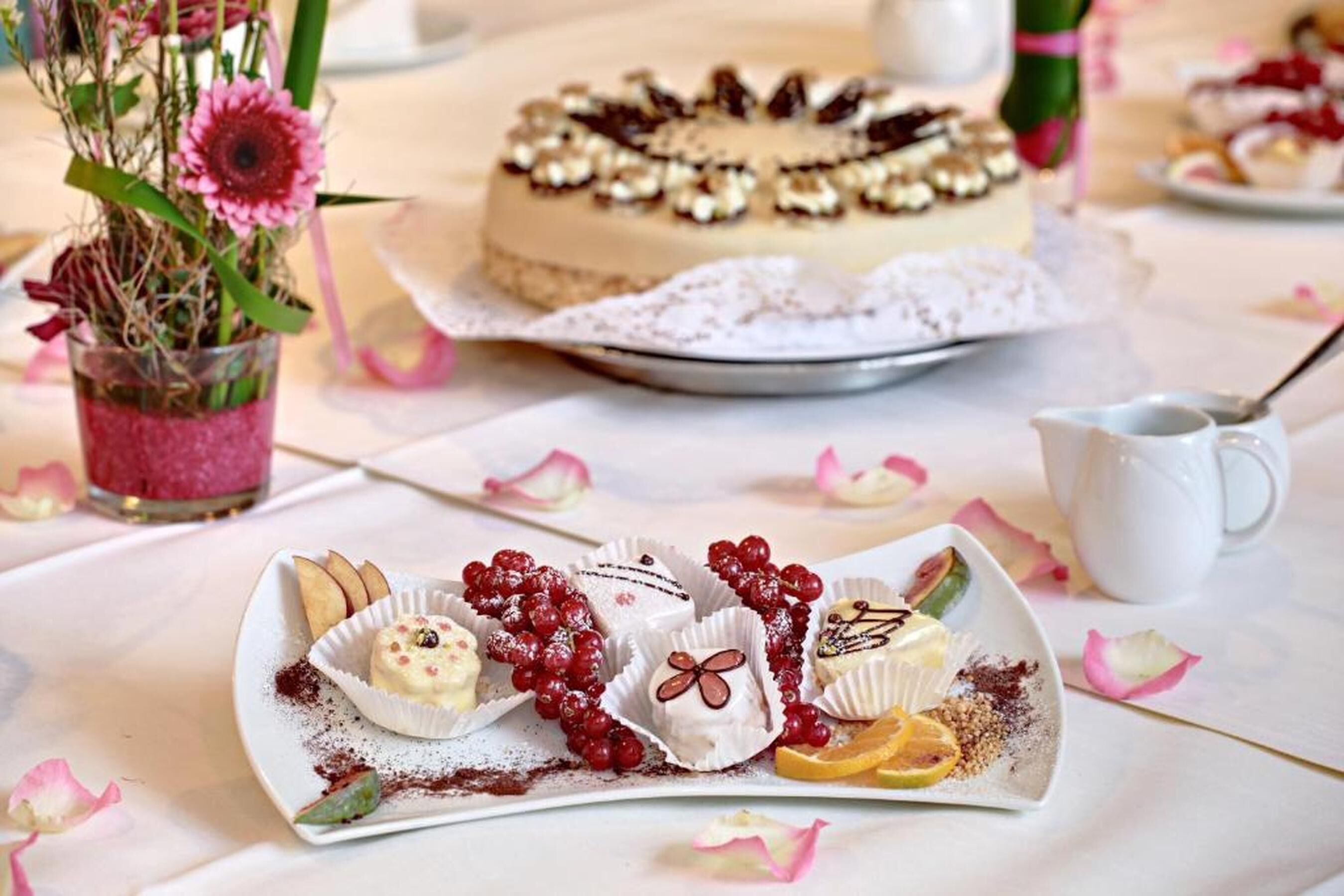A dessert table with various pastries and a cake, a glass vase with flowers, and a white pitcher on a table.