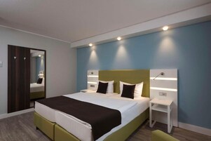 A modern hotel room with a large bed, bedside tables, a mirror, and a wardrobe.
