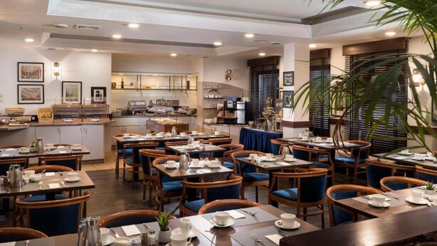 A hotel dining area with tables and chairs, a kitchen in the background, and a large plant. - Peine Salzgitter Hotel by Belvilla (Peine)