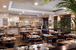 A hotel dining area with tables and chairs, a kitchen in the background, and a large plant.
