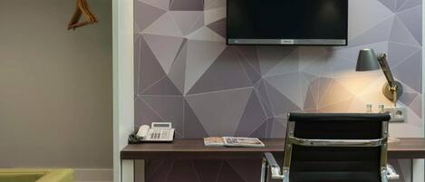 A modern office space with a flat-screen TV mounted on a geometric-patterned wall, a desk with a telephone and magazine, a black office chair, and a lamp.