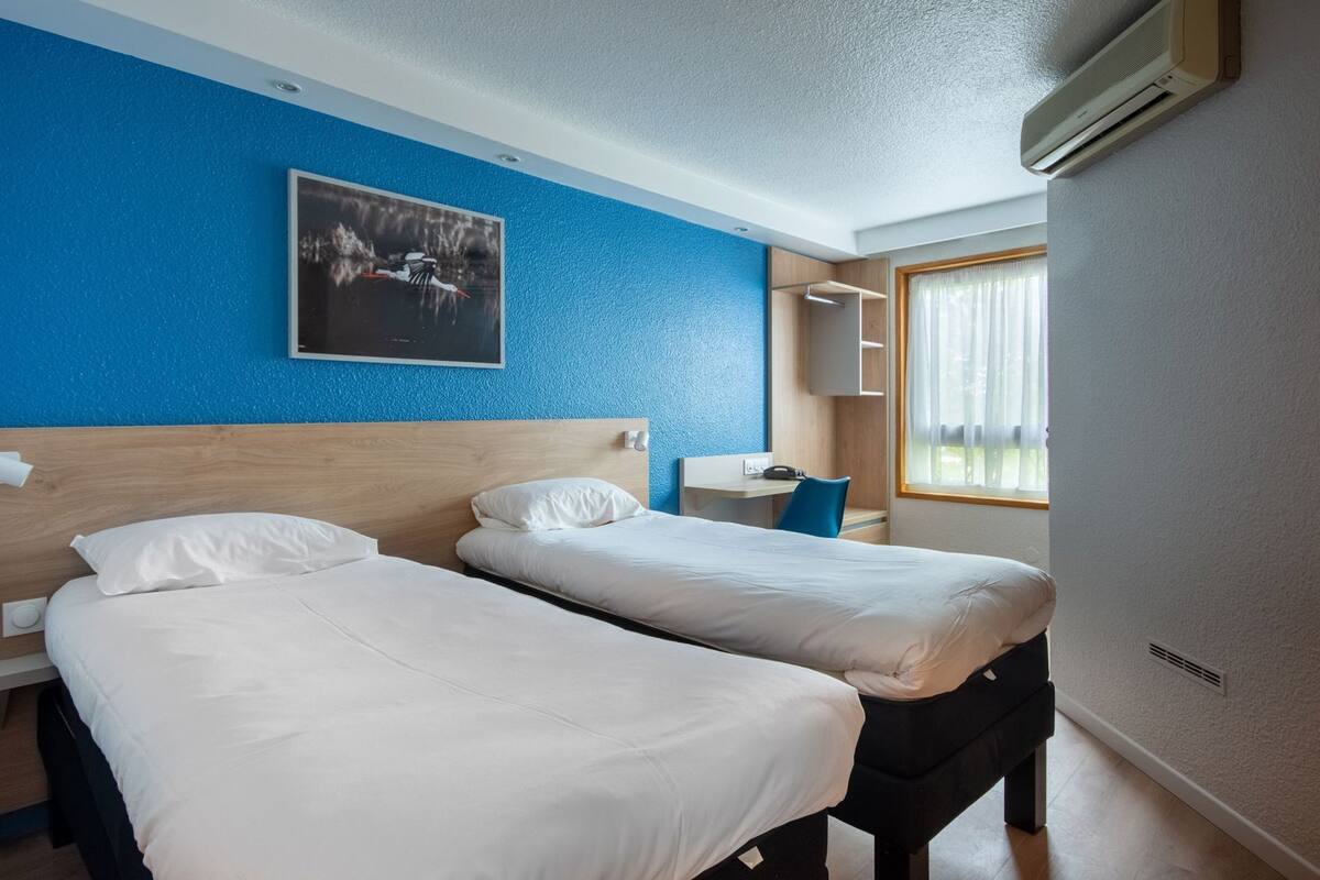 Une chambre d’hôtel avec deux lits, un bureau, une chaise, une fenêtre avec des rideaux et un climatiseur fixé au mur.