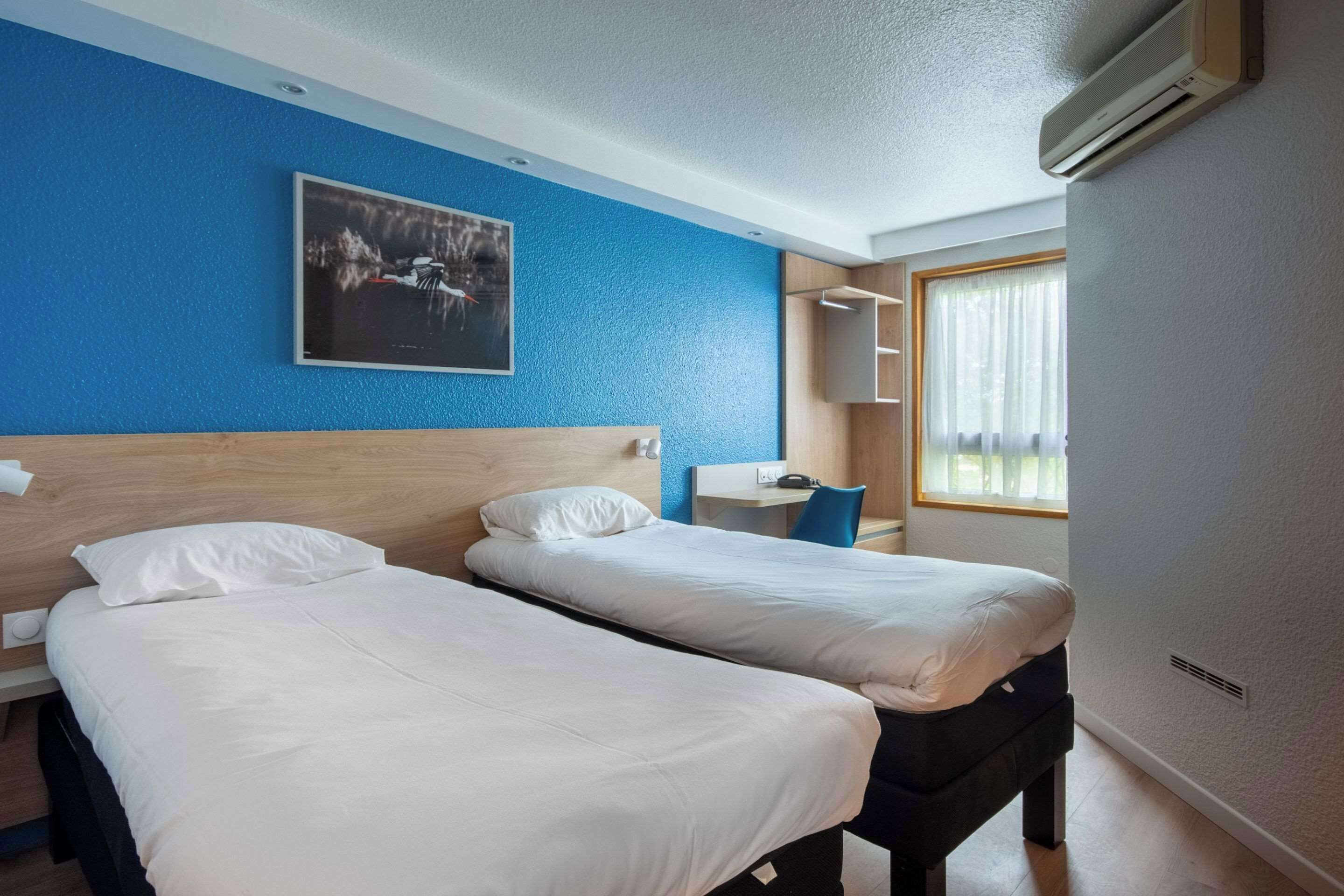 A hotel room with two single beds, a desk, a chair, a window with curtains, and a wall-mounted air conditioner.