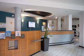 A hotel reception area with a wooden reception desk, a digital screen displaying the hotel name, and a potted plant. - Brit Hotel Confort Mulhouse Centre (Mulhouse)
