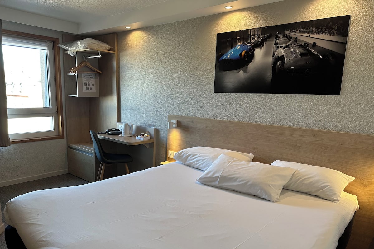 Ein Hotelzimmer mit Bett, Schreibtisch, Stuhl, Fenster und einem Wandbild mit Rennautos.
