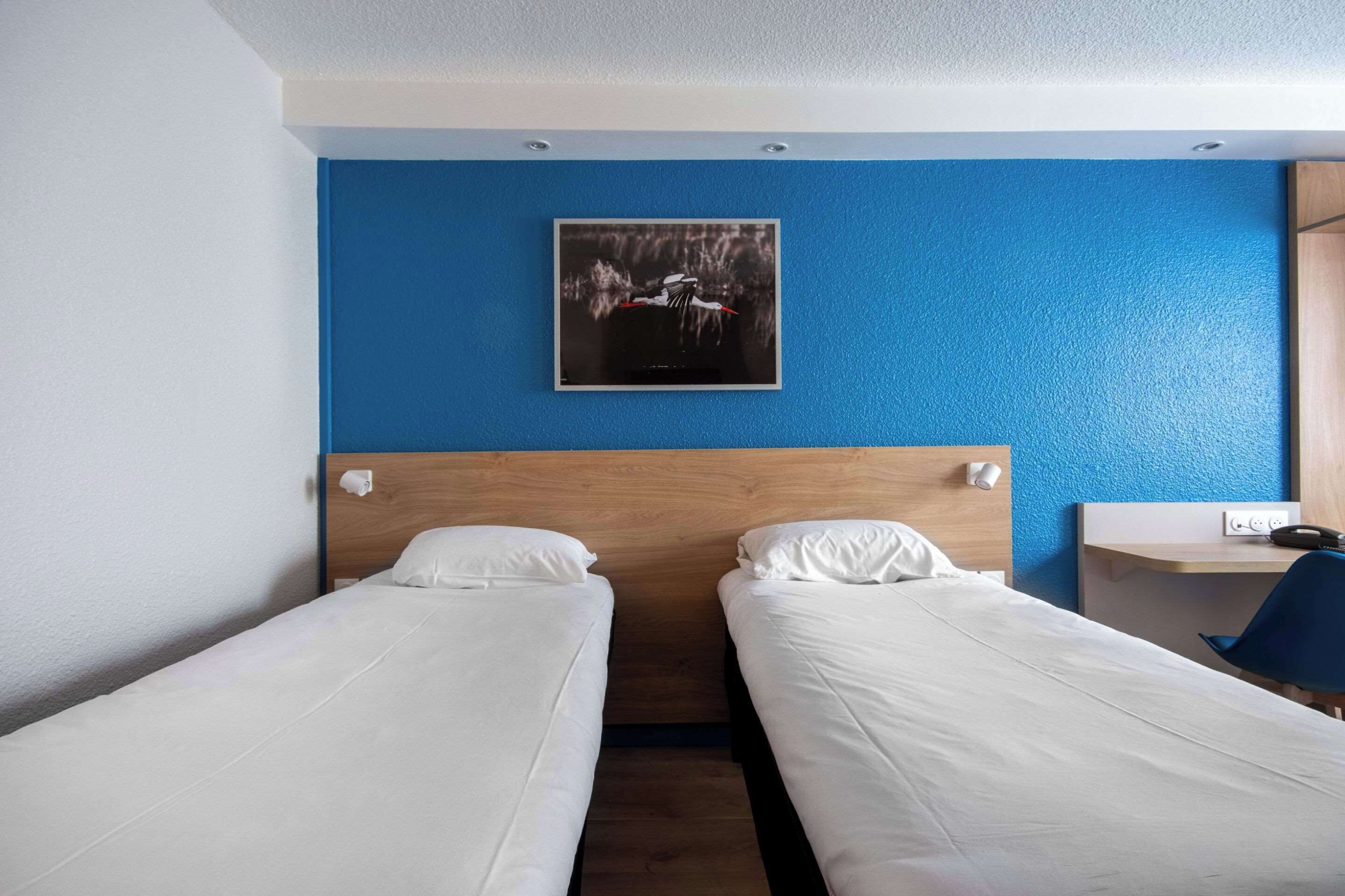 Ein Hotelzimmer mit zwei Einzelbetten, einem Holz-Kopfteil, einer blauen Akzentwand, einem gerahmten Bild und einem Schreibtisch mit Stuhl.