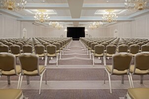 Un salón de conferencias con filas de sillas, un escenario, candelabros y suelo alfombrado.