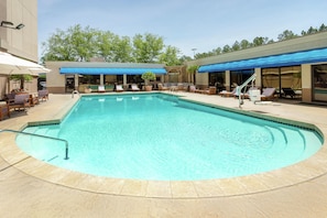 A swimming pool with lounge chairs and umbrellas, surrounded by a building with large windows and a covered patio area.
