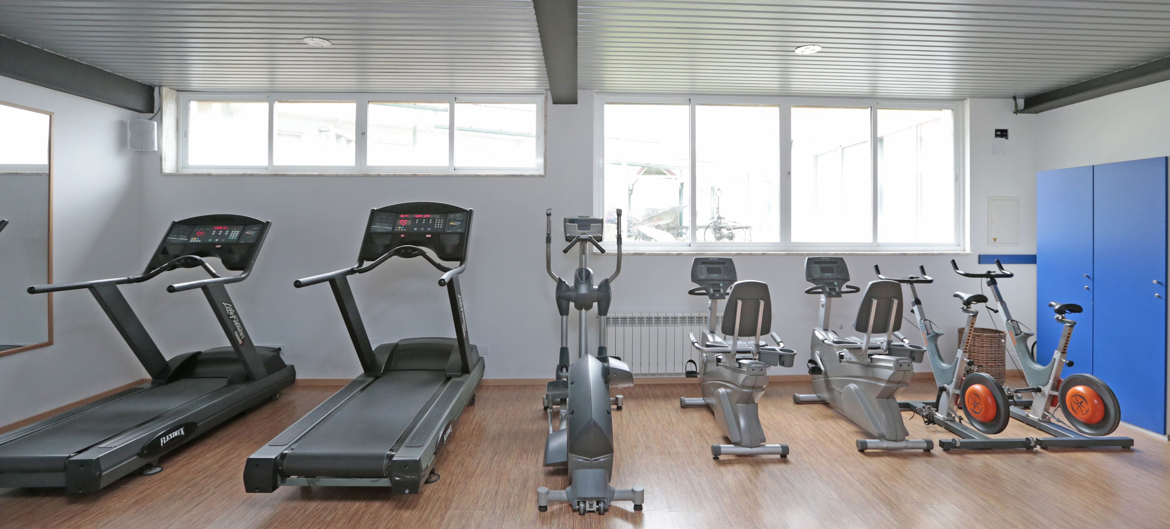 Un gimnasio equipado con cintas de correr, elípticas y bicicletas estáticas. Ventanas ofrecen luz natural y vistas al exterior.
