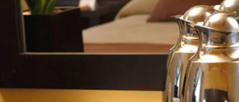 A hotel room with a bed, a bedside table with a coffee pot, cups, and pastries.