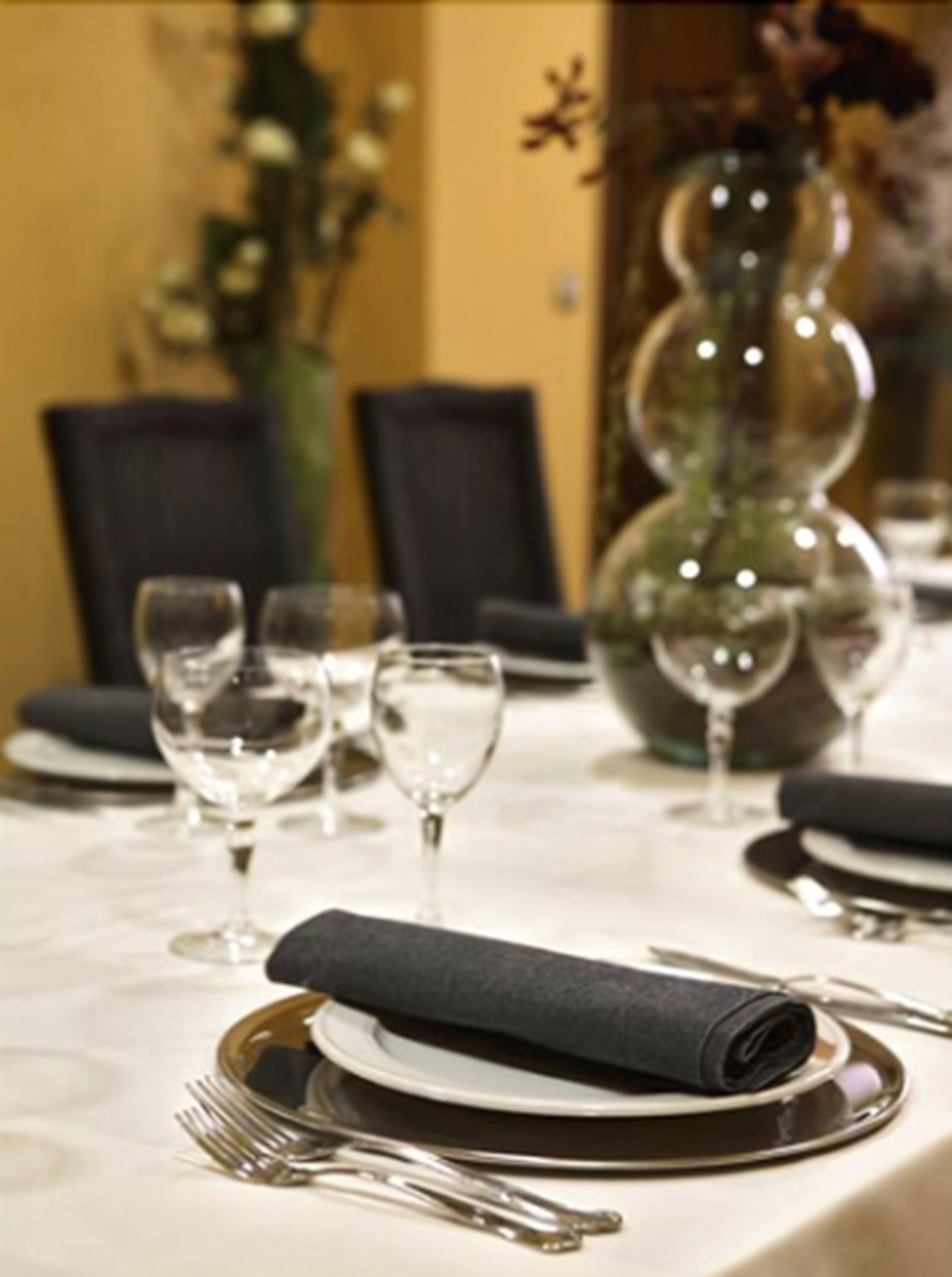 A dining table set with plates, cutlery, and wine glasses, with a vase of flowers in the background.