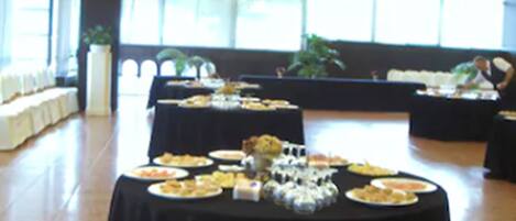 A spacious hall with a high ceiling and large windows, featuring a long table with various food items and a seating area with a sofa and chairs.